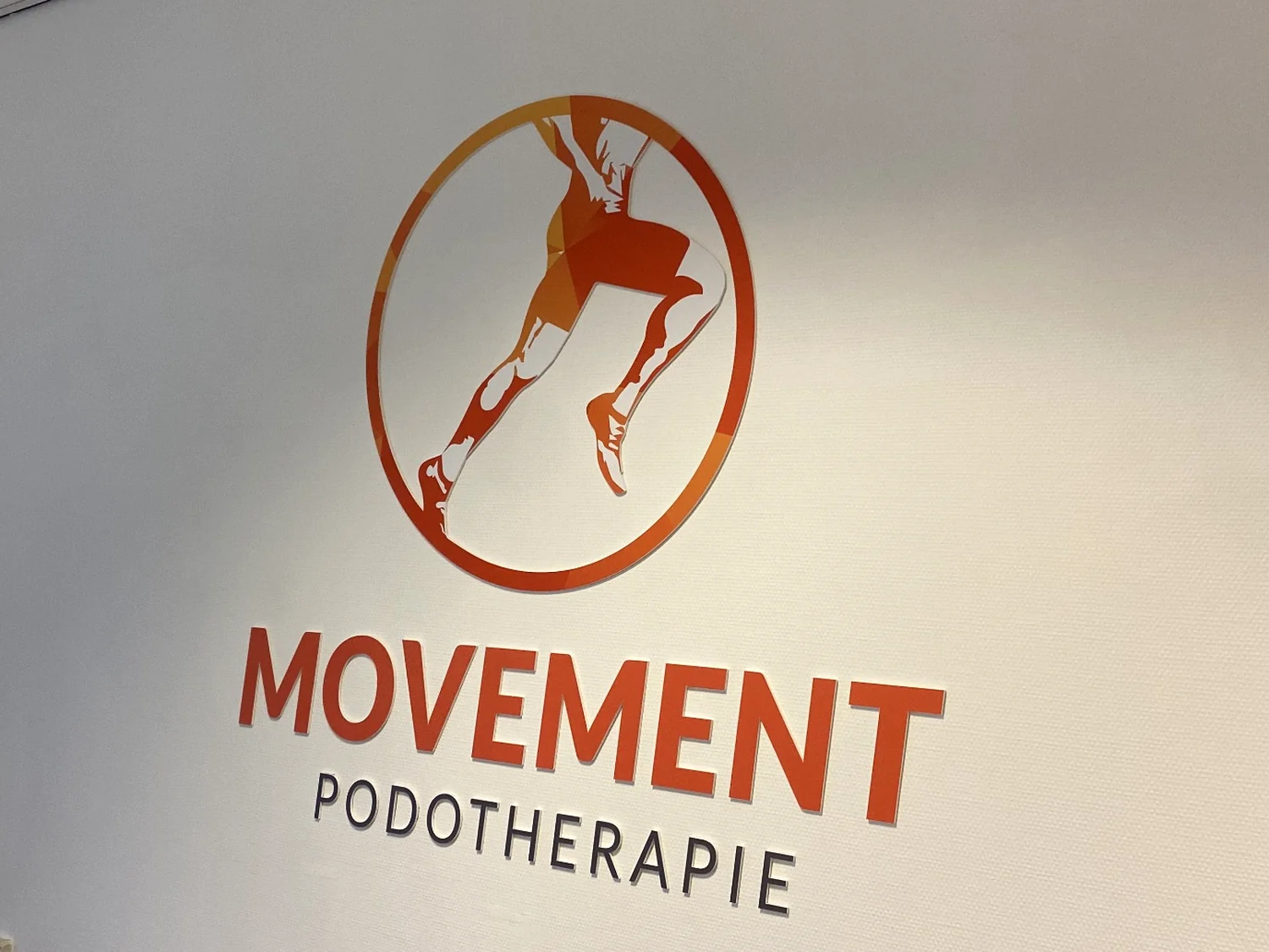 Wandlogo Movement Podotherapie in oranje en zwart, bedrukt op 5 mm wit PVC en gemonteerd aan de wand